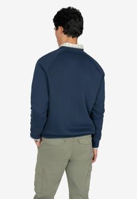 Marineblå sweatshirt med rund hals, blød tekstur og raglanærmer, båret over en plaidskjorte, parret med lys grøn cargo bukser.