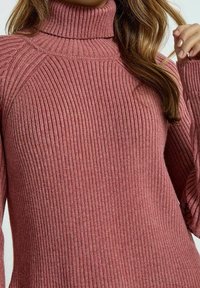 Pull à col roulé côtelé en rose poussiéreux, avec des manches longues et une coupe ajustée. Fabriqué en fil texturé doux pour la chaleur.