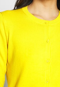 Gelber Strickcardigan mit runder Ausschnitt, der fünf Knöpfe auf der Vorderseite und ein strukturiertes Material aufweist.