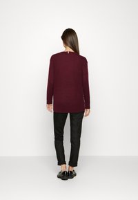Bordeaux lange mouwtrui met een ontspannen pasvorm, geribde manchetten en zoom, en een achterlabel met een rood, wit en blauw accent. In combinatie met zwarte broek.