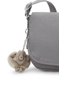 Bolso de hombro de tela gris con solapa, que cuenta con un pequeño llavero de un mono de peluche adjunto cerca del zipper.