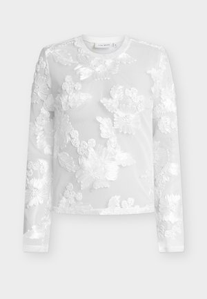 Haut blanc à manches longues en tissu transparent avec des motifs floraux brodés, col rond et petite étiquette de marque au niveau du col.
