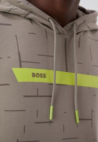 Närmast på en beige huvtröja med ett mönster av tunna mörka linjer, en neonfärgad horisontell rand och texten "BOSS" ovanför snöre ändarna med neonfärgade spetsar.