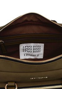Interno di una borsa verde oliva che mostra una fodera marrone, una tasca con cerniera e un'etichetta bianca con testo stampato: "DEVO RESISTERE... L'HO COMPRATA."