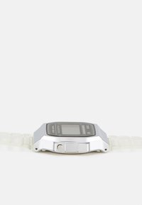 Montre numérique avec un boîtier métallique argenté, un affichage LCD noir et un bracelet en plastique transparent à maillons segmentés.