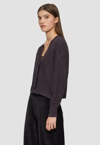 Lila Strickjacke mit tiefem V-Ausschnitt, langen Ärmeln und einem einzigen Knopfverschluss. Kombiniert mit lockeren schwarzen Cordhosen.