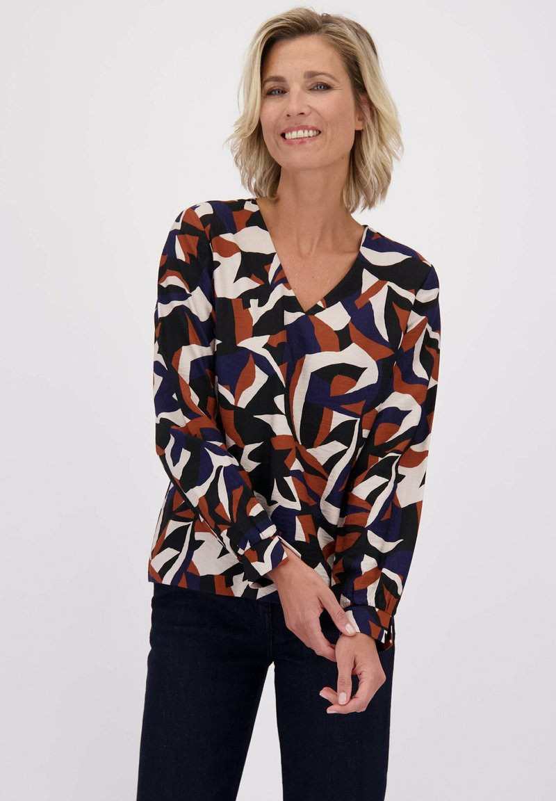 Vrouw die glimlacht, gekleed in een lange mouwtop met abstracte zwarte, witte, bruine en marineblauwe vormen, en donkere broek tegen een effen achtergrond.