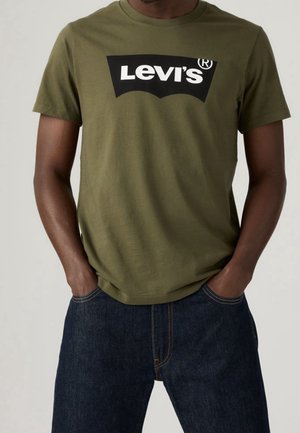 Mies, joka pukeutunut oliivinvihreään Levi's-t-paitaan, jossa mustavalkoinen logo, tummansiniset farkut, kädet taskuissa, seisoo yksinkertaisen taustan edessä.