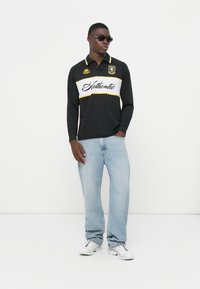 Polo shirt nero a maniche lunghe con una striscia orizzontale bianca e dettagli gialli, abbinato a jeans azzurri chiari e sneakers bianche.