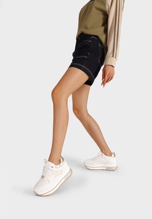 Sneakers bianchi con suola spessa e dettagli dorati, abbinati a pantaloni corti di denim scuro. I pantaloni corti hanno cuciture visibili e un design aderente.