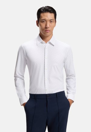 Homme portant une chemise blanche à manches longues avec un petit motif à pois et un pantalon bleu foncé, mains dans les poches, expression neutre.