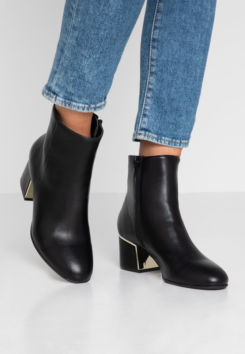 Bottes basses en cuir noir avec talons blocs et accents dorés réfléchissants, portées avec un jean bleu coupé au niveau des chevilles.