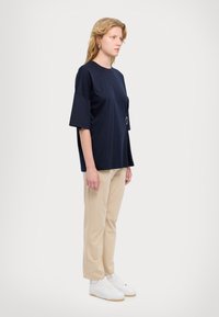Mørkeblå oversized t-skjorte med korte ermer, med en stor frontlomme og en sølvfarget sirkulær detalj, sammen med beige bukser og hvite sneakers.