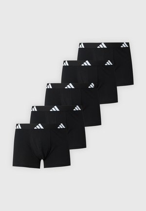 Homme torse nu debout pieds nus portant un boxer noir avec un logo blanc sur la ceinture, sur un fond blanc uni.