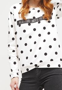 Biała bluza z czarnymi kropkami, ozdobiona poziomym czarnym paskiem z napisem „ALL YOU NEED IS LOVE”. Ściągacze na mankietach i u dołu bluzy.