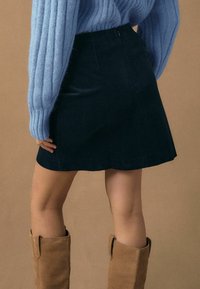 Mini-jupe en velours côtelé noir de forme évasée, associée à un pull côtelé bleu clair et des bottes hautes couleur noisette, sur un fond neutre.