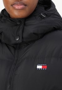 Primo piano di una persona che indossa un giubbotto imbottito nero con zip sul davanti e il logo Tommy Jeans sul petto.