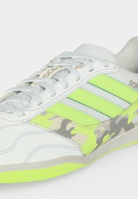Άσπρο αθλητικό παπούτσι με φωσφορίζουσες πράσινες ρίγες, γκρι και μπεζ καμουφλάζ λεπτομέρειες, άσπρα κορδόνια και ανοιχτό πράσινη σόλα.