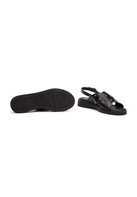 Sandalias de cuero negro con diseño recortado, tachuelas circulares y correa de talón. Suela plana con patrón de agarre texturizado.