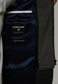 Szary wnętrze kurtki z granatową satynową podszewką, z czarnymi metkami z napisem "Strellson" i "Najlepsza Tkanina Stretch". Obecna kieszeń na zamek.