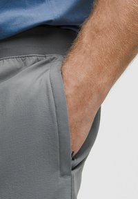 Grå sportsshorts med et struktureret stof og elastisk talje, der har en sidelomme til højre. Hånd hviler i lommen.