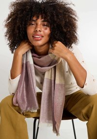 Écharpe légère dans des teintes de rose poudré, beige et argent, avec une texture douce et des bords frangés. Portée lâchement autour du cou.