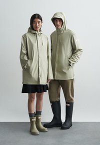 Beżowe wodoodporne kurtki z kapturem i zatrzaskami. Modele noszą khaki szorty i czarne kalosze, stojąc na szarym tle.