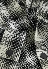 Tissu à carreaux noir et blanc avec des boutons-pression noirs visibles et des sections pliées, mettant en valeur la texture et les détails des boutons sur le vêtement.