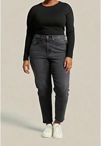 Personne portant un haut à manches longues noir, un jean gris foncé taille haute et des baskets blanches, debout devant un fond clair uni.