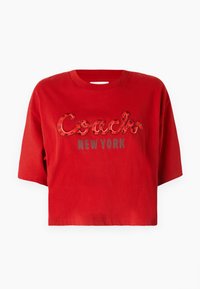 Rotes, kurzes Baumwoll-T-Shirt mit einem glänzenden, geschwungenen "Coach"-Logo in Rot, das ein kleines Schlangen-Muster aufweist, und grauem "NEW YORK"-Text darunter.