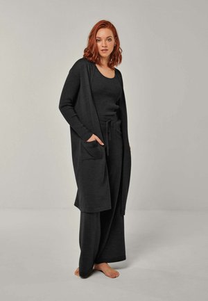 Frau mit roten Haaren, die eine schwarze lange Strickjacke, ein schwarzes Top und weite Hosen trägt, steht barfuß vor einem schlichten hellen Hintergrund.