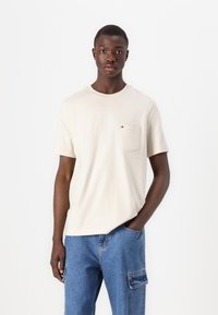 Beige crew-neck t-shirt made of cotton, featuring a front pocket and a small logo patch. Paired with blue cargo jeans.