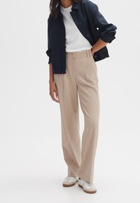 Beige, hooggerande broeken met plooien, gecombineerd met een wit T-shirt en een zwarte, cropped jas. De schoenen zijn wit met beige accenten.