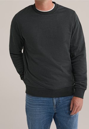 Mand iført ensfarvet mørkegrå langærmet sweatshirt og blå jeans mod ensartet lys baggrund, stående med den ene hånd i lommen.