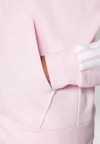 Světle růžová mikina se klokaní kapsou, pružnými manžetami a třemi bílými pruhy na rukávu. Měkká textura, ležérní design.