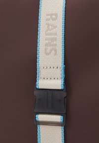 Beige verstellbarer Riemen mit hellblauen Nähten, ausgestattet mit einer schwarzen Kunststoffschnalle. Text ist auf der Oberfläche des Riemens geprägt.