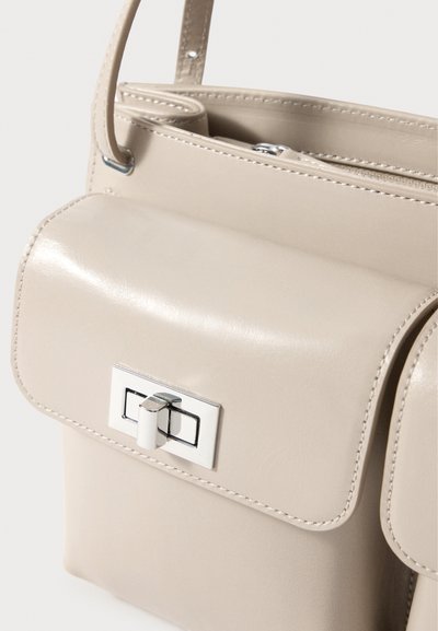 Bež nahkakott, millel on klappkinnitus, hõbedased detailid ja nähtavad õmblusjooned. Kompaktne disain ja struktureeritud kuju.