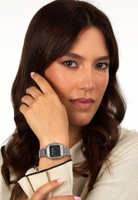 Silberne Uhr mit achteckigem Zifferblatt, schwarzem Ziffernblatt und klaren Edelsteinakzenten, getragen von einer Person mit welligem Haar, die sich mit einer Hand ins Gesicht greift.