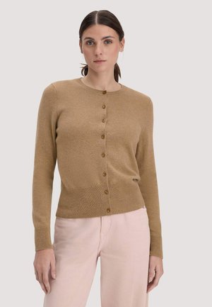 Vrouw met een beige, dichtgeknoopte cardigan en lichtroze broek, staand tegen een effen lichtgrijze achtergrond.