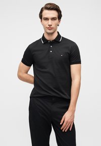 Μαύρο πόλο πουκάμισο με λευκή επένδυση γιακά, κοντά μανίκια και τρία κουμπιά, με μικρό λογότυπο στο αριστερό μέρος του στήθους.