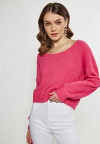 Maglione tricot in fucsia con scollatura ampia e maniche lunghe, abbinato a jeans bianchi aderenti, esposto su uno sfondo neutro.
