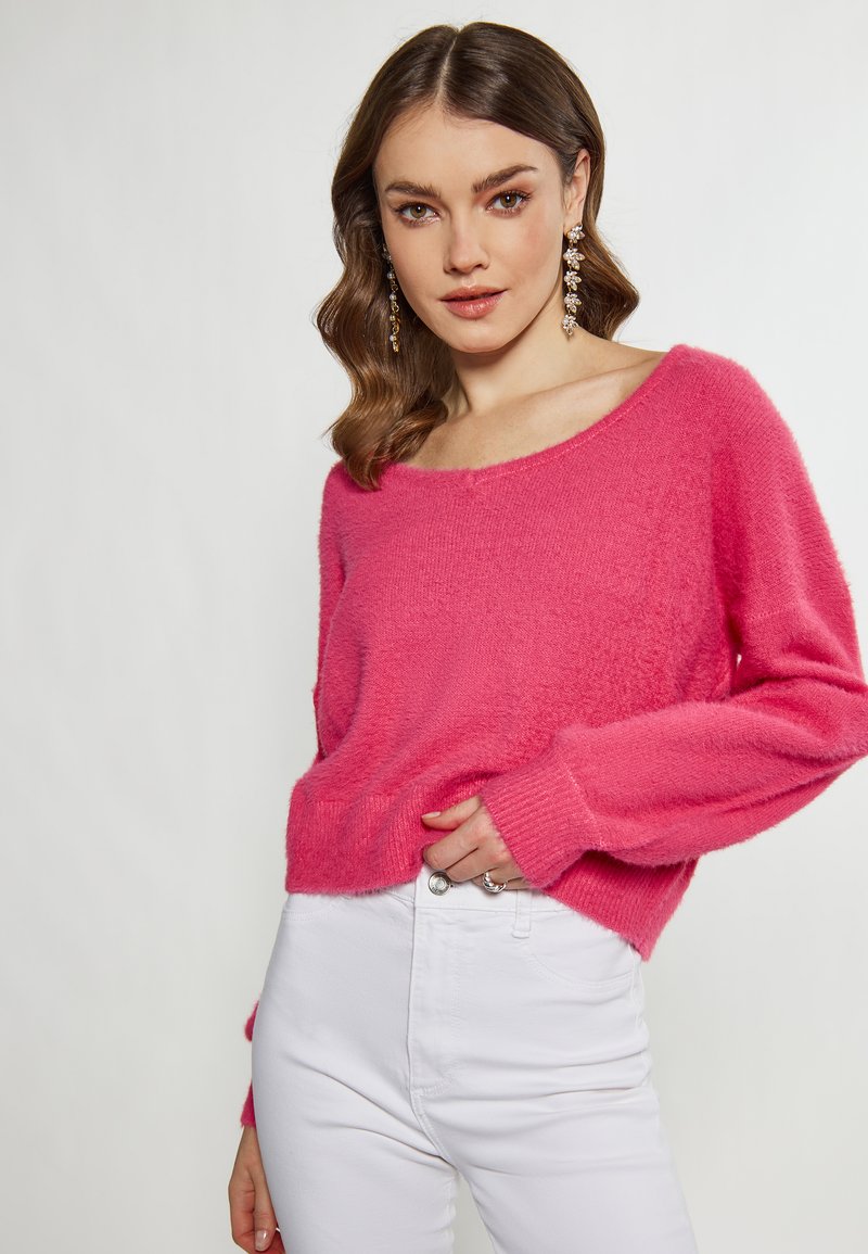 Maglione tricot in fucsia con scollatura ampia e maniche lunghe, abbinato a jeans bianchi aderenti, esposto su uno sfondo neutro.