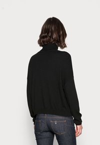 Donna con capelli castani alla spalla, che indossa un maglione nero a collo alto e jeans blu scuro, di spalle su uno sfondo chiaro e uniforme.