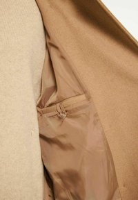 Brunt ydre stof med en lysere beige inderside. Har en glat tekstur, en knapdetalje og en lomme indeni.