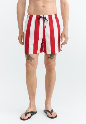 Mann trägt rot-weiß vertikal gestreifte Badeshorts mit schwarzer Kordel, schwarzen Flip-Flops und sichtbaren Bein-Tattoos vor einfachem Hintergrund.
