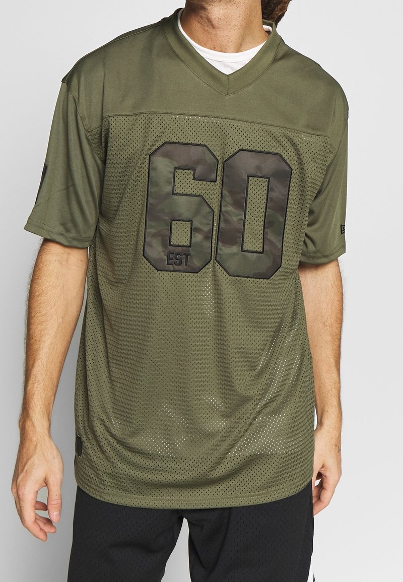 Man i olivgrön mesh sporttröja med stort kamouflagemönstrat nummer "60 EST" över en vit t-shirt och svarta shorts, stående mot en enfärgad bakgrund.