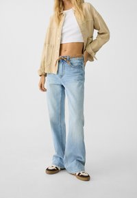 Veste en denim oversized beige clair, top court blanc, jeans loose bleu clair et baskets marron avec des accents blancs. Tenue décontractée et textures douces.