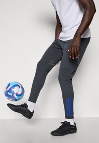 Grå atletiske bukser med slim fit, med blå striber og en gylden detalje. Båret med sorte fodboldstøvler. En fodbold ligger i nærheden.