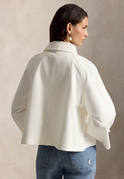 Γυναίκα που φορά χαλαρό λευκό σακάκι με φαρδιά μανίκια και μπλε τζιν, εμφανιζόμενη από πίσω σε ουδέτερο φόντο.
