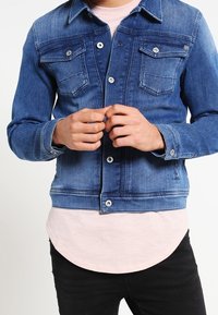 Chaqueta de mezclilla en azul medio con dos bolsillos en el pecho, cierres de botones y costuras en contraste. Llevada sobre una camisa rosa claro con dobladillo curvado.